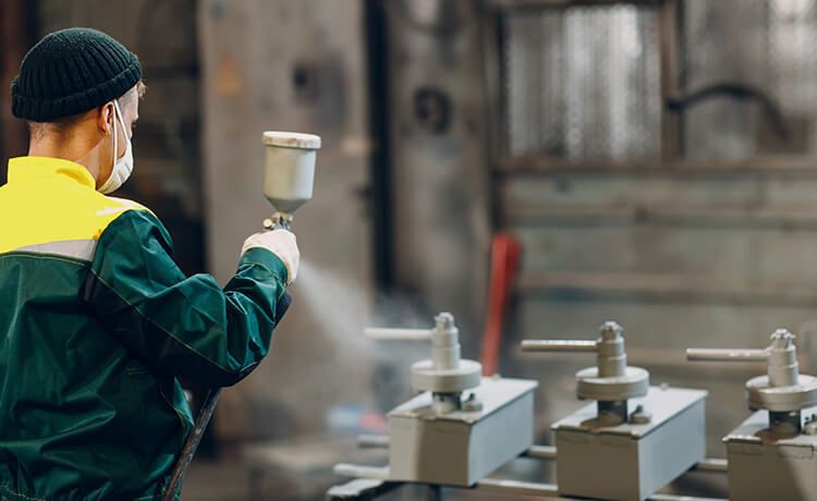 Oberflächenbeschichter-Azubi besprüht Metallteile mit einer Pulvergrundierung.